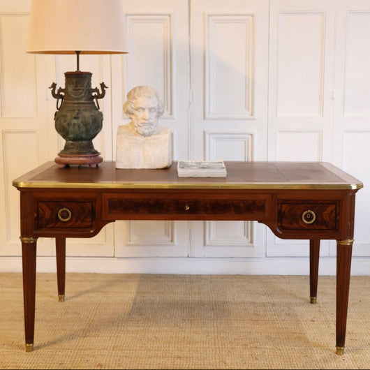 AN EARLY 20TH CENTURY FRENCH MAHOGANY DESK WITH REEDED LEGS, FLAME MAHOGANY VENEERED PANELS, BRASS MOUNTS AND ORIGINAL LEATHER TOP.
