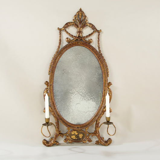 An elegant Adam period oval girandole or dressing mirror, the gilt carved wood and composition frame with an anthemion crest, two flanking candle arms and a panel of Chinese lacquer at the base. English, circa 1770.