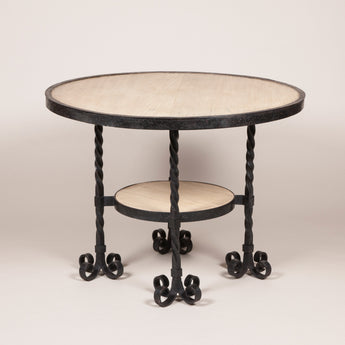 A round wrought iron table, the top and small round under-tier inset with bleached mahogany and the four legs each with four scrolled feet. Probably French, mid-20th century.